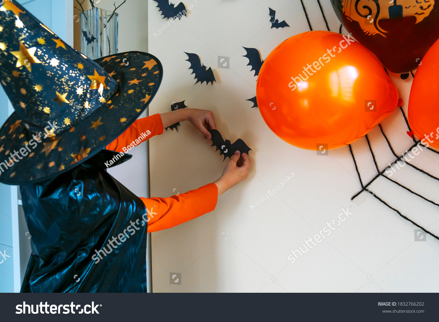 Boy in a black cape and a witch's hat glues a paper bat to the wall. The child decorates the house for Halloween. #1832766202