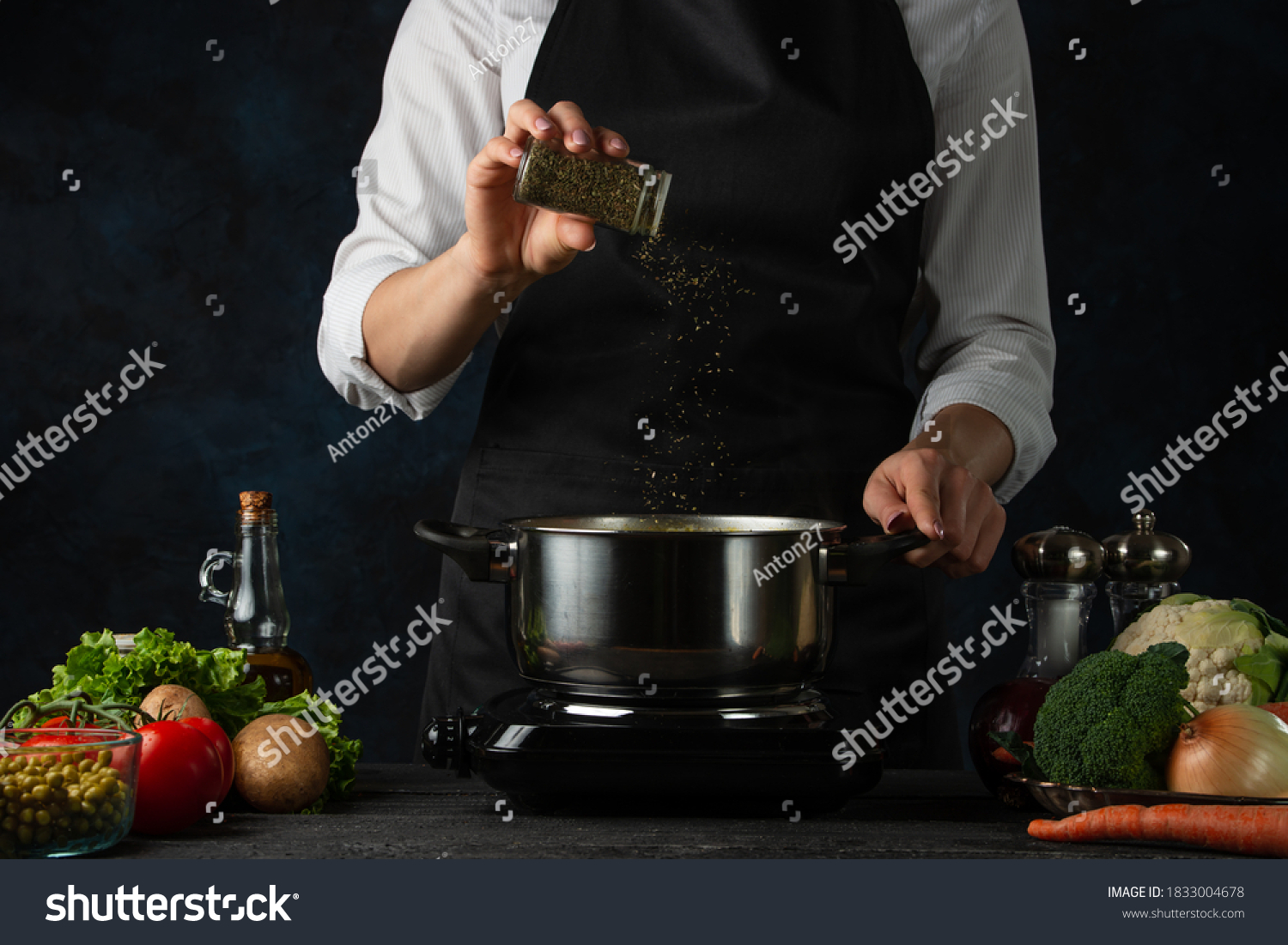 The professional chef in black apron adds spices into pot with soup on dark blue background. Backstage of cooking dinner. Preparing meal concept. Frozen motion. #1833004678