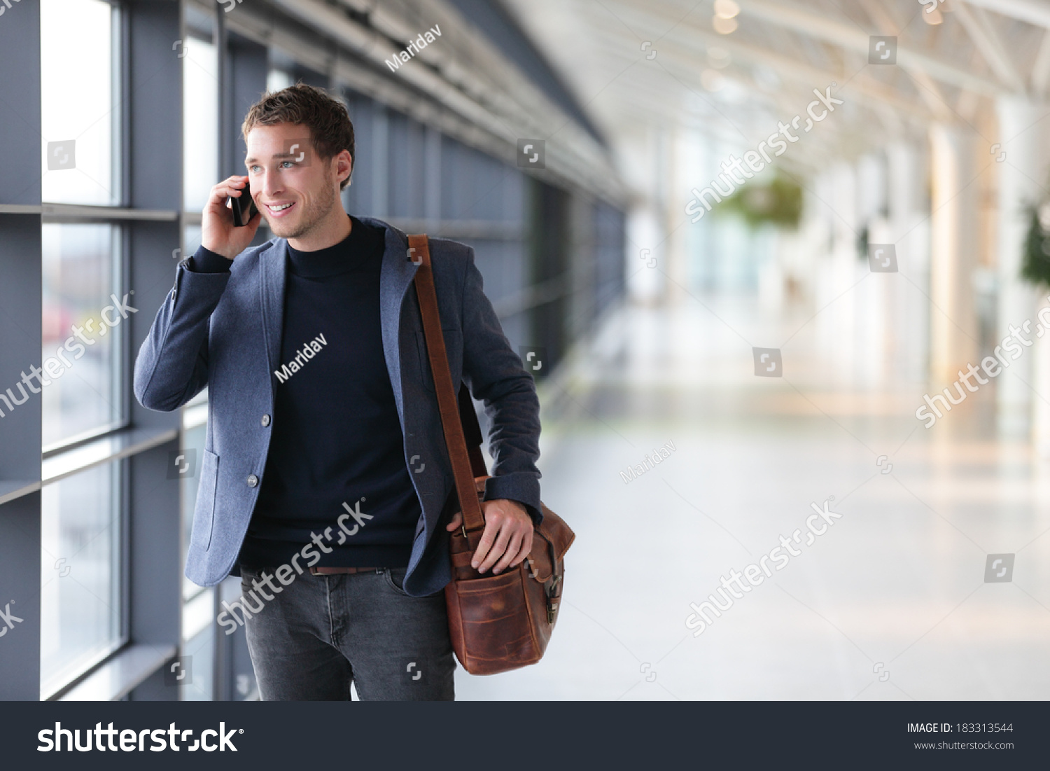 Urban business man talking on smart phone traveling walking inside in airport. Casual young businessman wearing suit jacket and shoulder bag. Handsome male model in his 20s. #183313544
