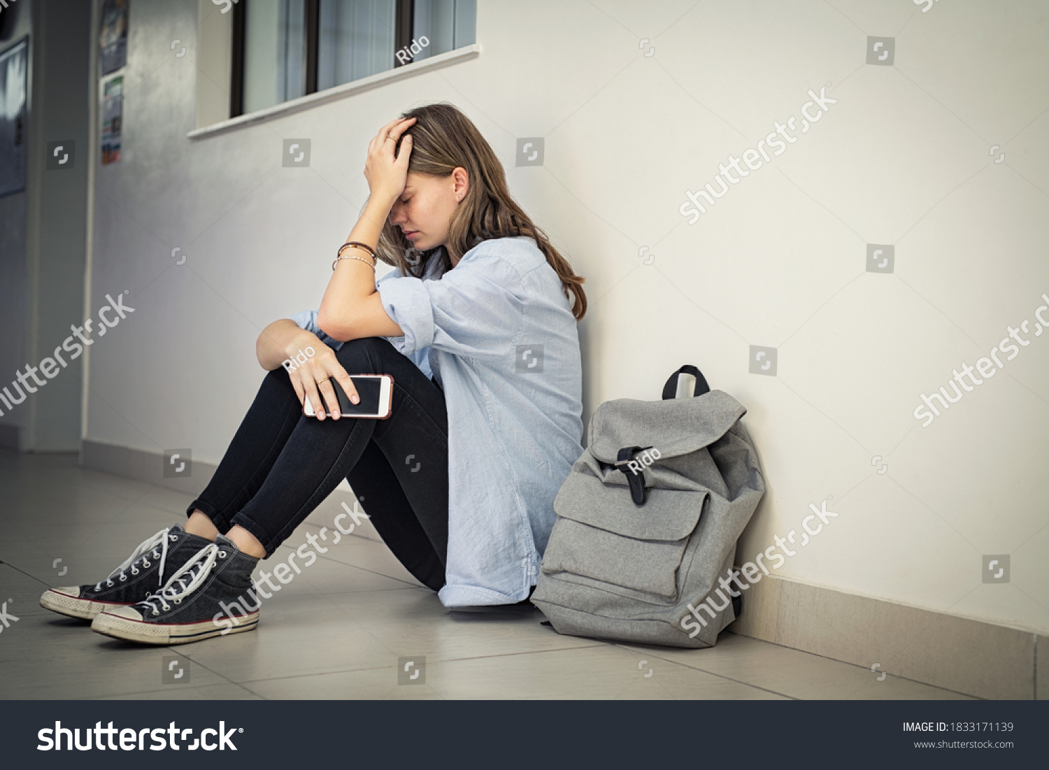 Upset and depressed girl holding smartphone sitting on college campus floor holding head. University sad student suffering from depression sitting on floor at high school. Lonely bullied teen. #1833171139