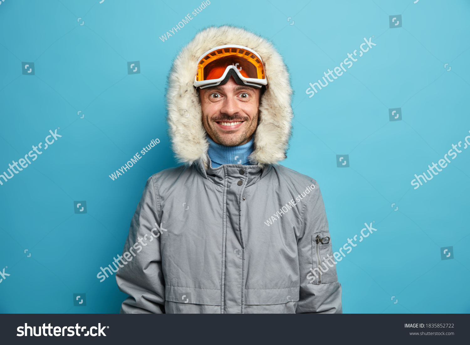 Sporty glad man enjoys winter sport recreation smiles gladfully wears ski goggles and grey jacket isolated on blue. Holidays tour recreation and traveling concept. Snowboarder leads active lifestyle #1835852722