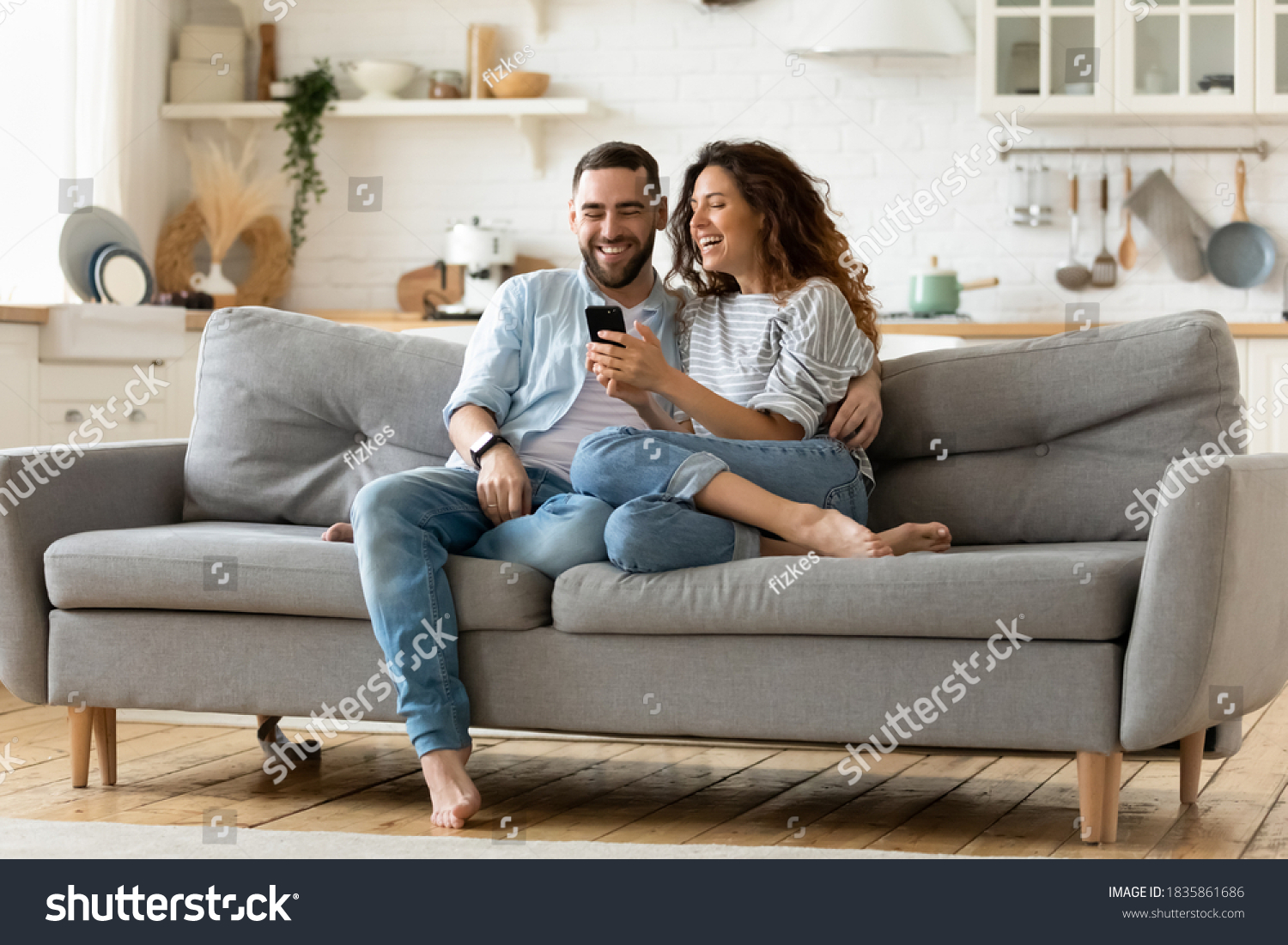Happy young woman and man hugging, using smartphone together, sitting on cozy couch at home, smiling overjoyed wife and husband looking at phone screen, sitting on sofa in modern living room #1835861686