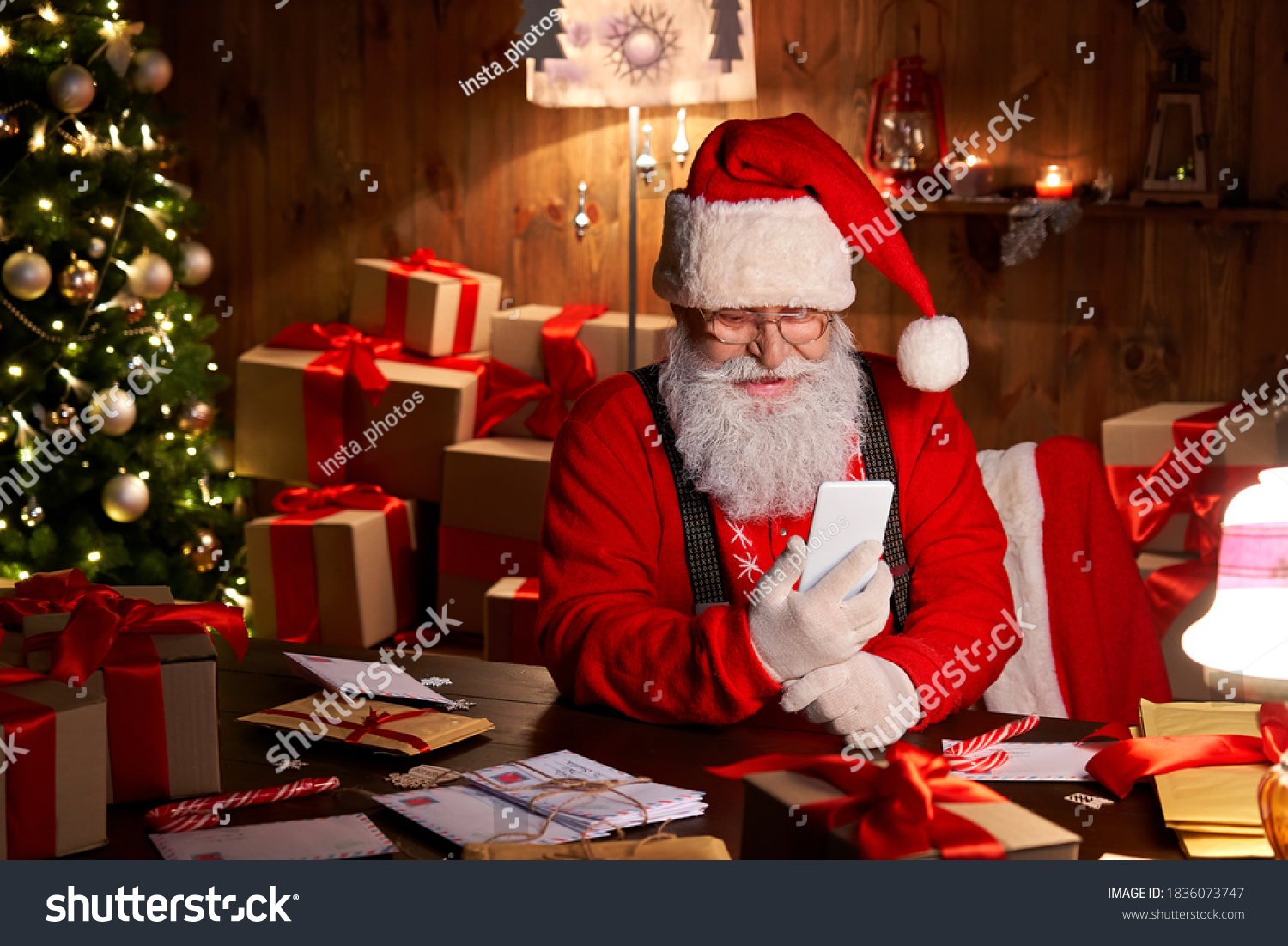 Happy Santa Claus holding smartphone holding cell phone using mobile app for social distance communication, watching videos or video calling in virtual online chat sit at home table late on xmas eve. #1836073747