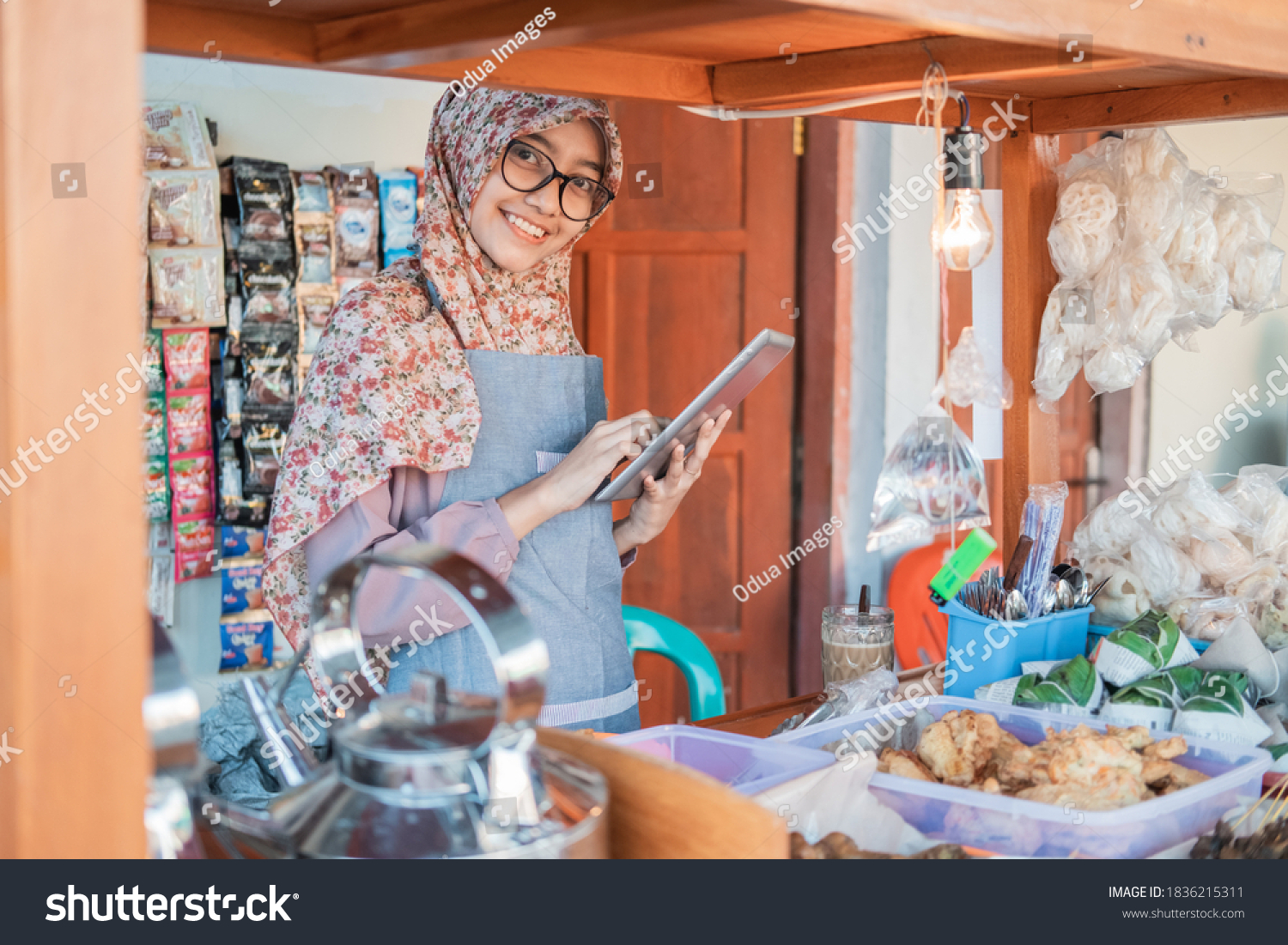 Asian female angkringan seller with hijab and apron using a tablet pc standing in side of the cart stall #1836215311