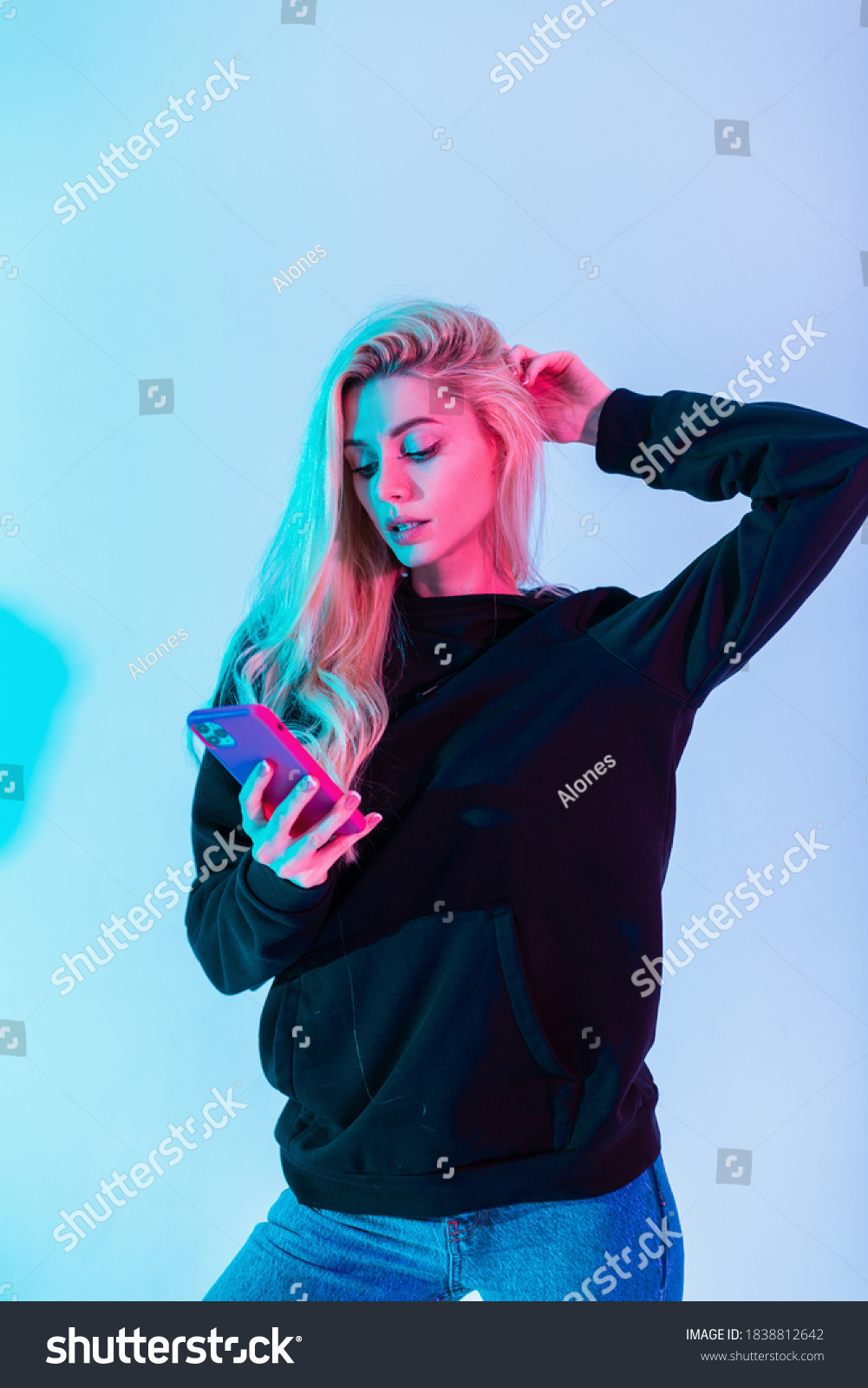 Glamorous beautiful woman model with pretty beauty face in fashion black hoodie and blue jeans with a phone on a colorful neon pink light background #1838812642