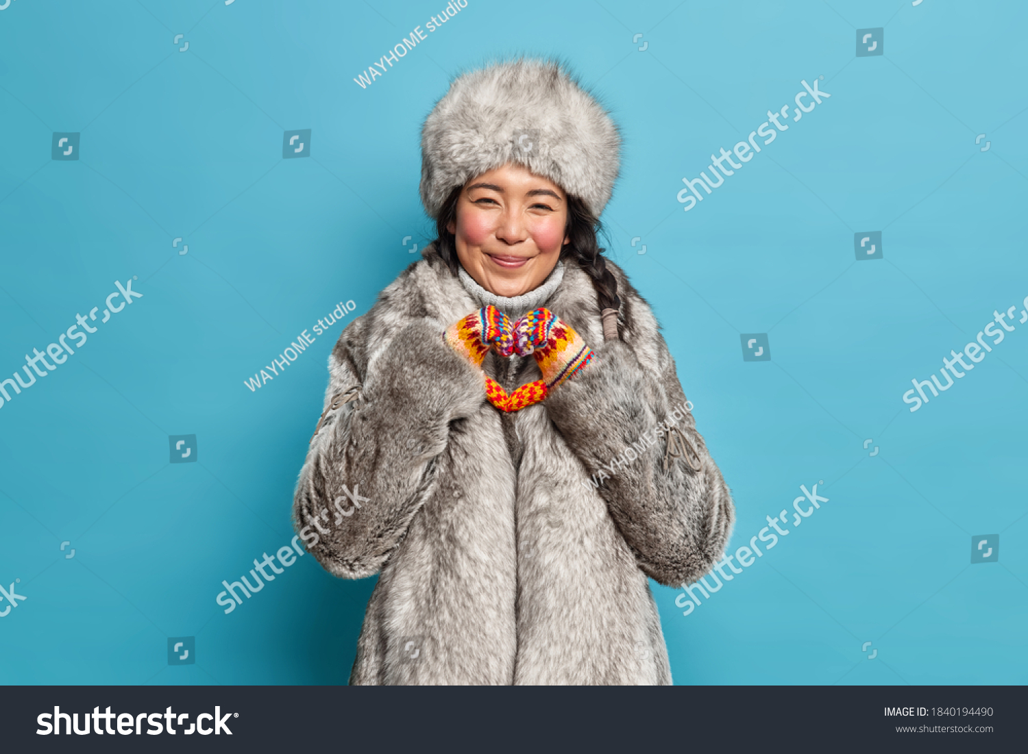 Cheerful eskimo woman shapes heart gesture expresses love dressed in warm winter clothing isolated over blue background. Affectionate reindeer herder in fur outerwear. Nothern people and cold season #1840194490