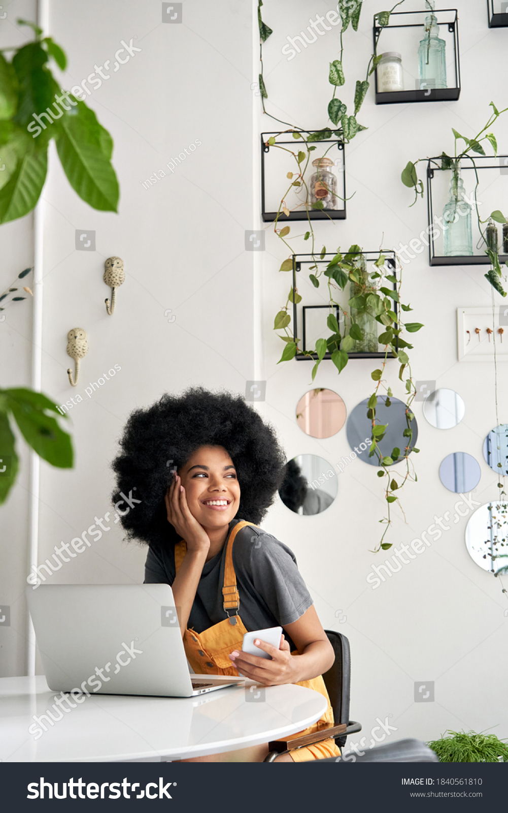 Young happy stylish African American gen z hipster teenage girl student with Afro hair laughing, looking through window, sitting at table in modern cozy cafe interior, holding phone, using laptop. #1840561810