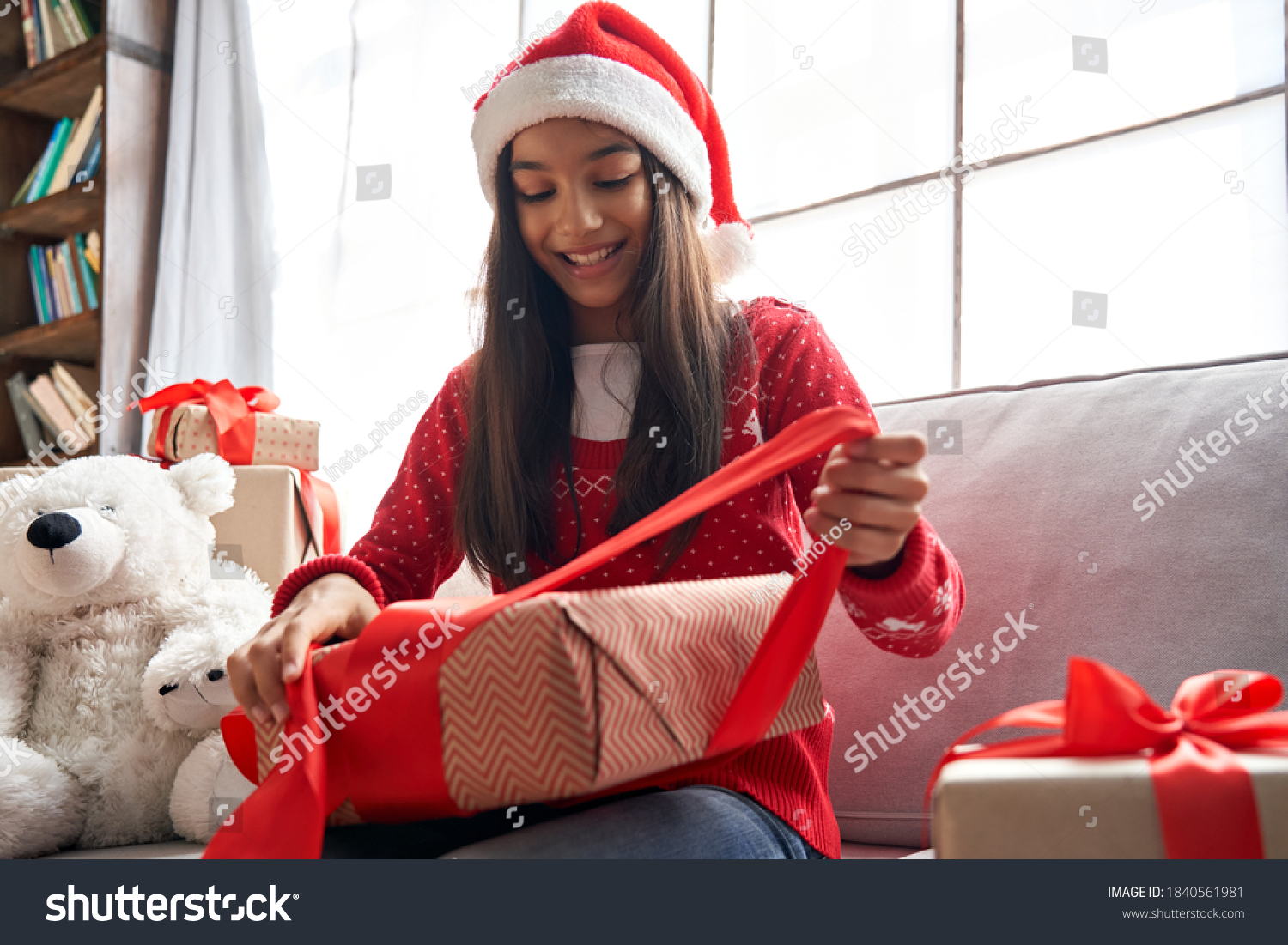 Happy indian kid girl unties red ribbon opens Christmas gift box at home. Smiling cute latin child wearing santa hat receiving xmas New Year present sitting on couch celebrating winter holidays. #1840561981