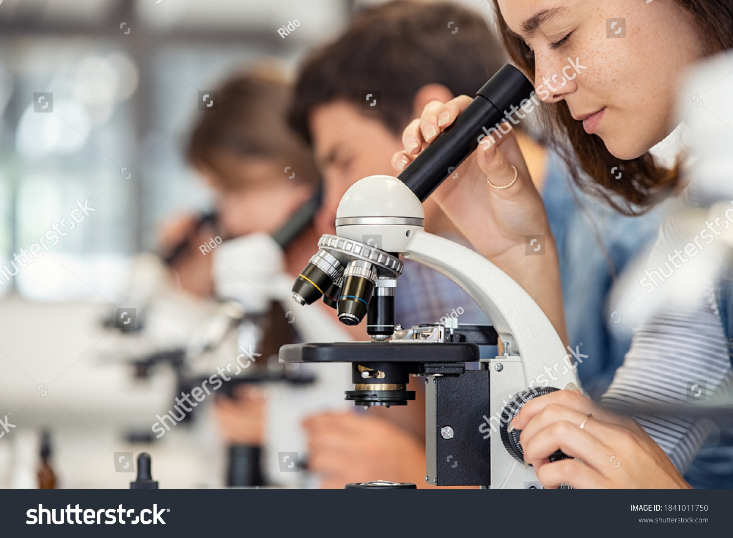 Close up of young woman seeing through microscope in science laboratory with other students. Focused college student using microscope in the chemistry lab during biology lesson. #1841011750