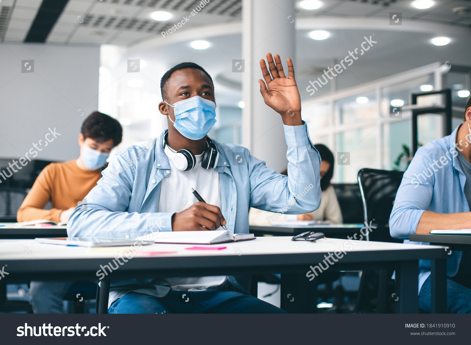 Education, Pandemic And Health Concept. African american male student raising hand for answer, wearing protective medical mask for protection from virus disease at high school lesson. New Normal #1841910910