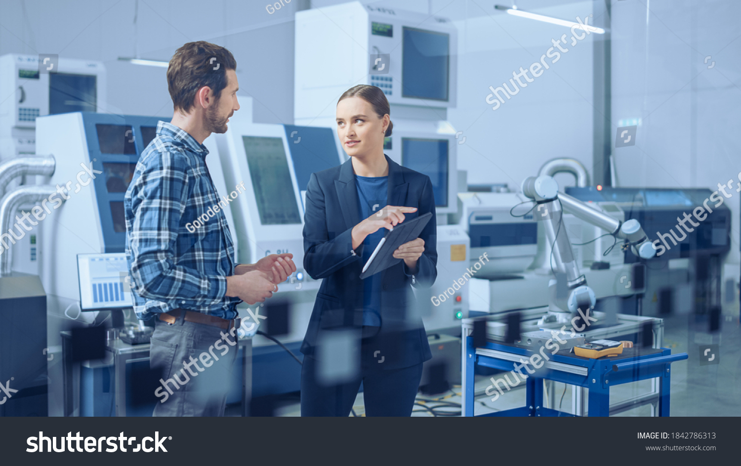 Modern Factory: Female Project Manager and Male Engineer Standing, Talking, Using Digital Tablet for Programming and Monitoring Assembly Line. Industry 4.0 Facility with High-Tech CNC Machinery #1842786313