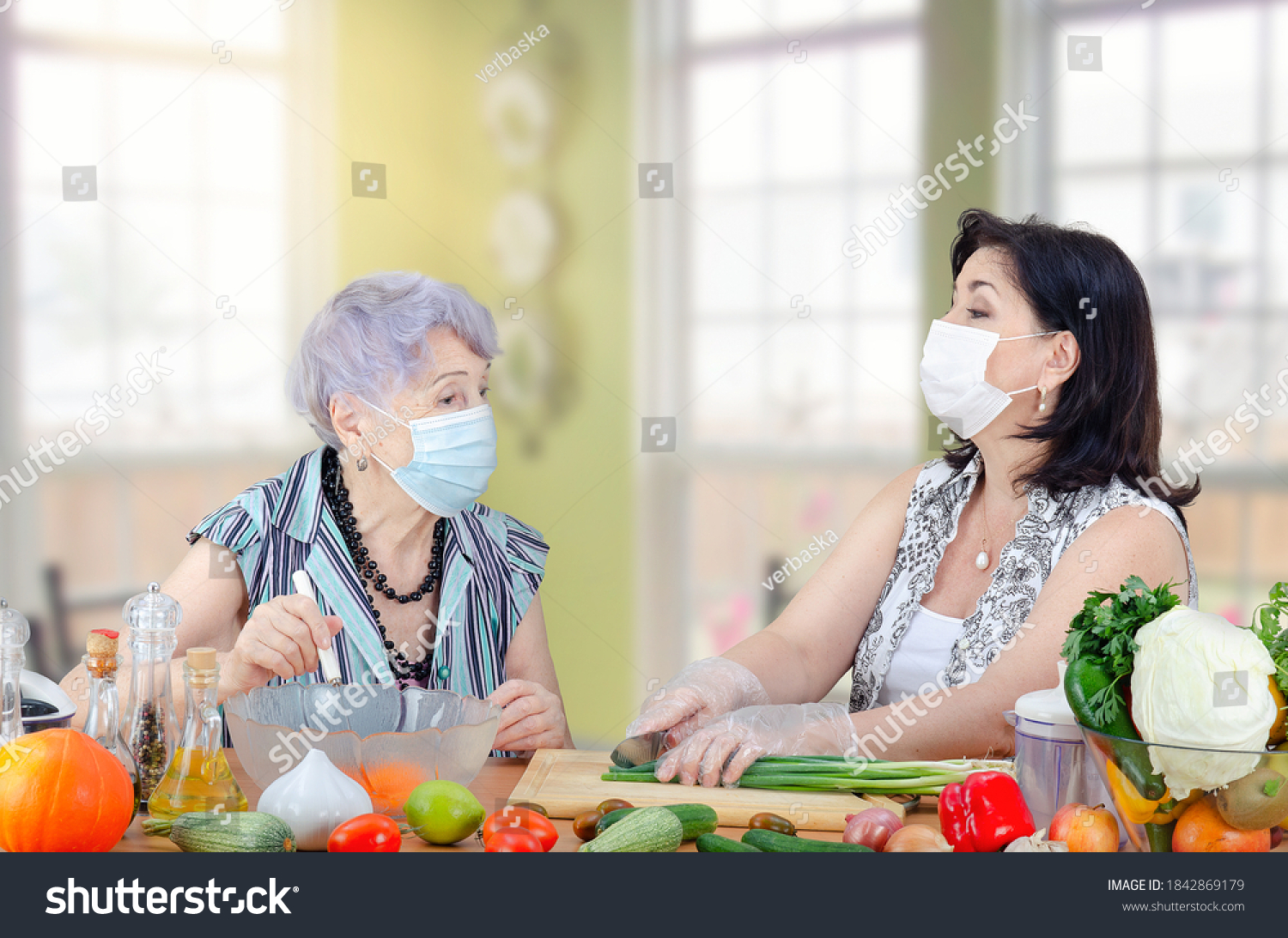 Caregiver or companion and senior adult woman speak in a friendly manner as they cook a vegetable salad together. Both are wearing protective face masks due to the coronavirus pandemic now.  #1842869179