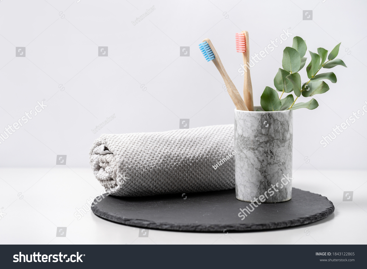 Concept of biodegradable objects. Two bamboo toothbrush in cup near bathroom towel and eucalyptus plant on black plate stand against white copy space background. Oral and dental care #1843122865