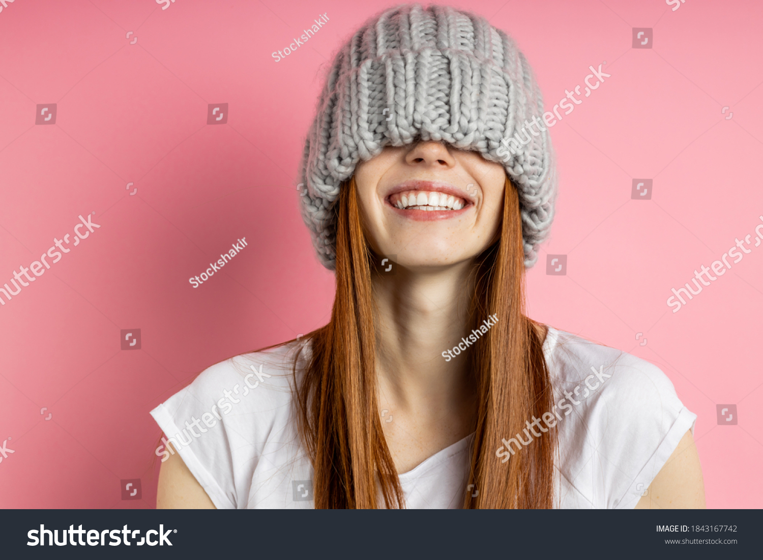 Close up portrait of cheerful happy girl with red long hair having fun, covering eyes with big loop knitted hat, smiling broadly with perfect white teeth isolated over pink background. #1843167742