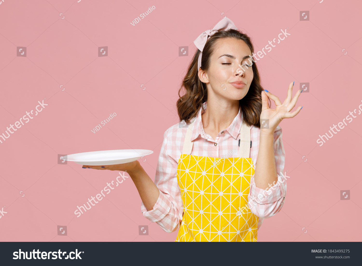 Cute young woman housewife in yellow apron hold empty plate making okay taste delight sign keeping eyes closed doing housework isolated on pastel pink background studio portrait. Housekeeping concept #1843499275