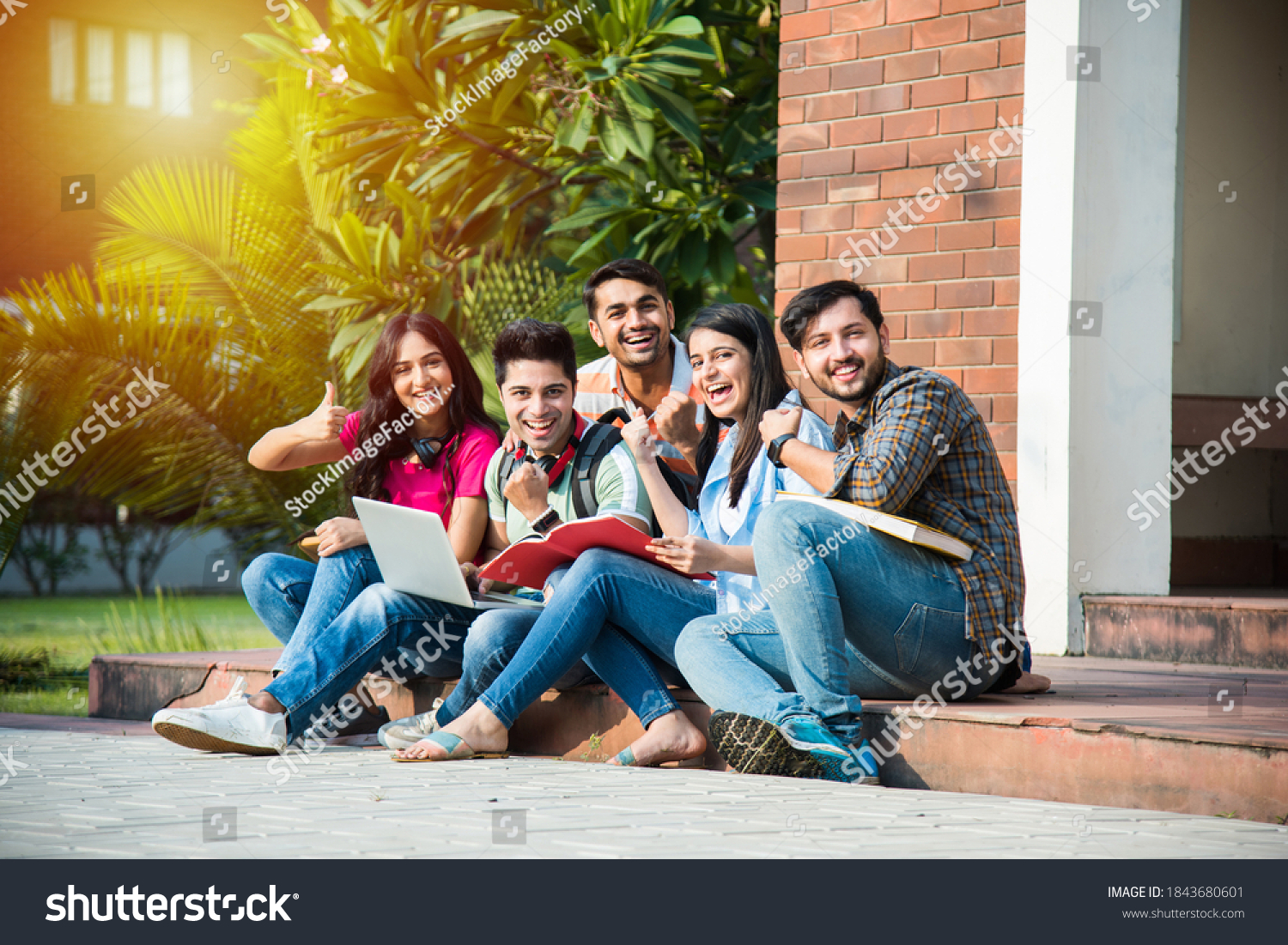 Young Asian Indian college students reading books, studying on laptop, preparing for exam or working on group project while sitting on grass, staircase or steps of college campus #1843680601