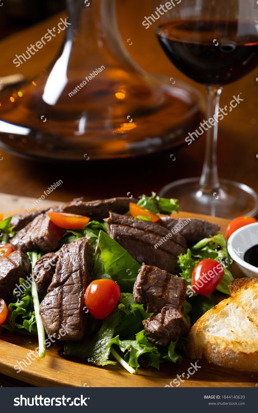 close up of grilled beef tenderloin fillet with rocket salad and cherry tomatoes pairing with Italian fine red wine bottle, glass and decanter in a wooden table classic vintage elegant moody style #1844140639