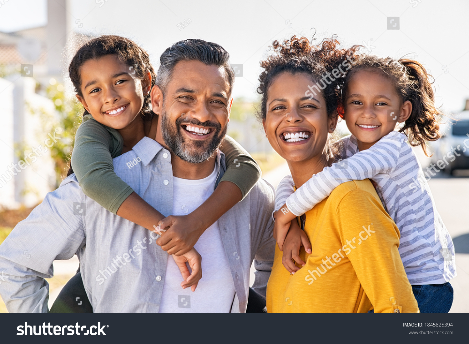 Mixed race parents giving piggyback ride to their children. Portrait of happy african mother and indian father with daughters looking at camera. Smiling family standing with their little girls. #1845825394