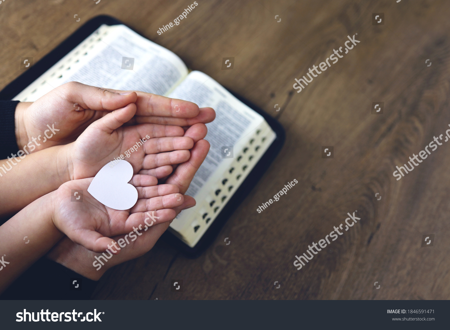 Religious Christian girl praying with her mother indoors. Bible in background. Hands holding white paper heart. Space for text #1846591471