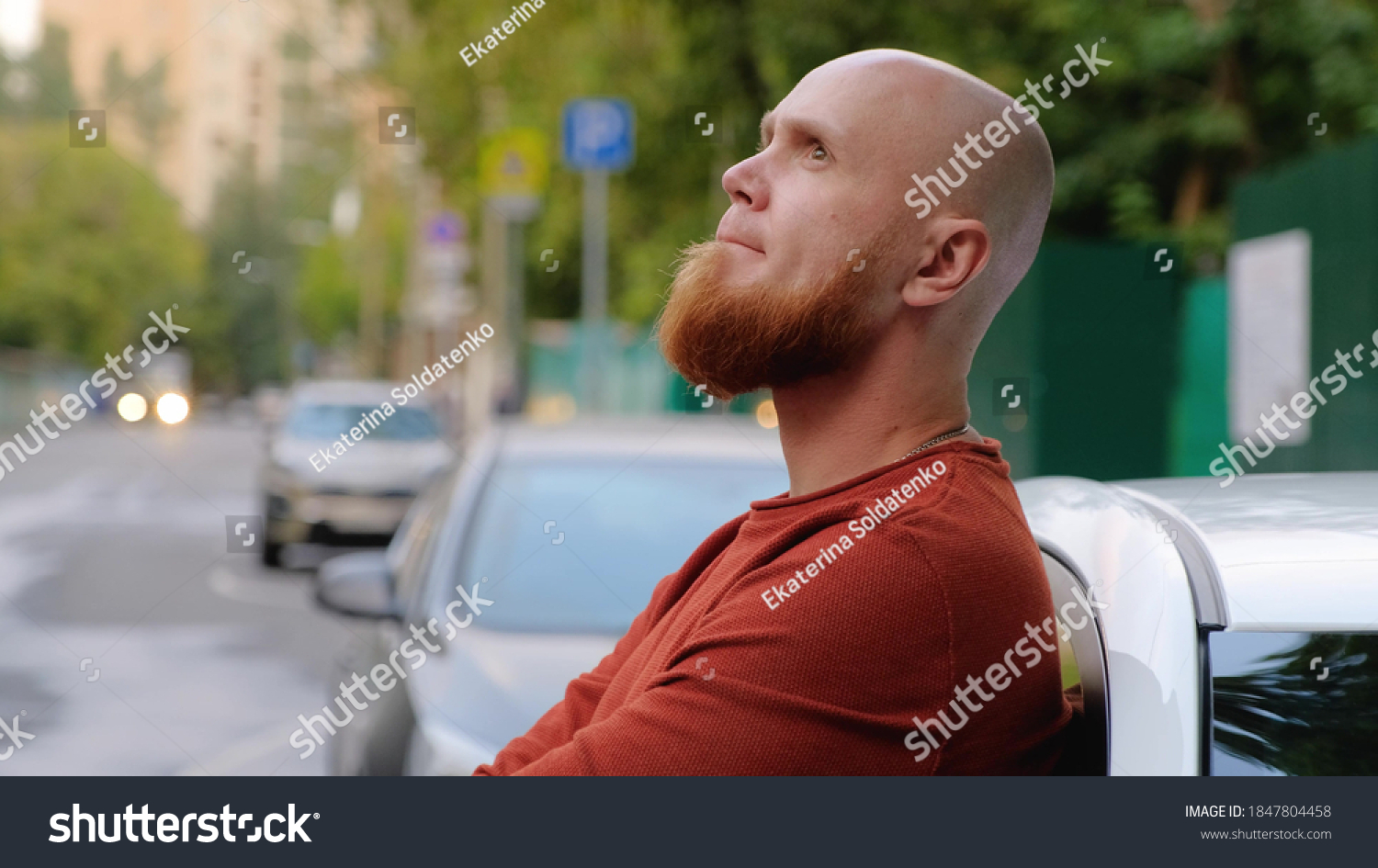 A young brutal bald guy with a well-groomed red beard and a sweater stands in the street, leaning against a car in a pensive state. Reflections on life. Summer evening. #1847804458