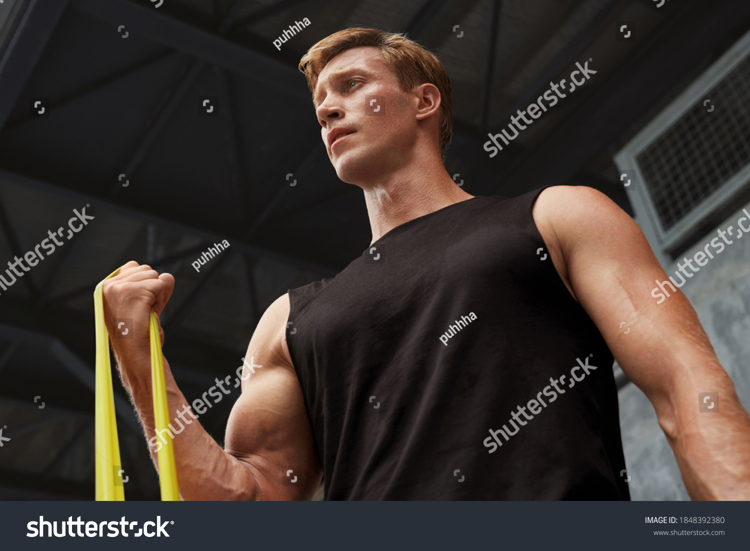 Young Man Stretching With Resistance Band Against Concrete Wall Outdoors. Handsome Caucasian Sportsman With Strong Muscular Body In Fashion Sportswear Doing Stretch Workout. #1848392380