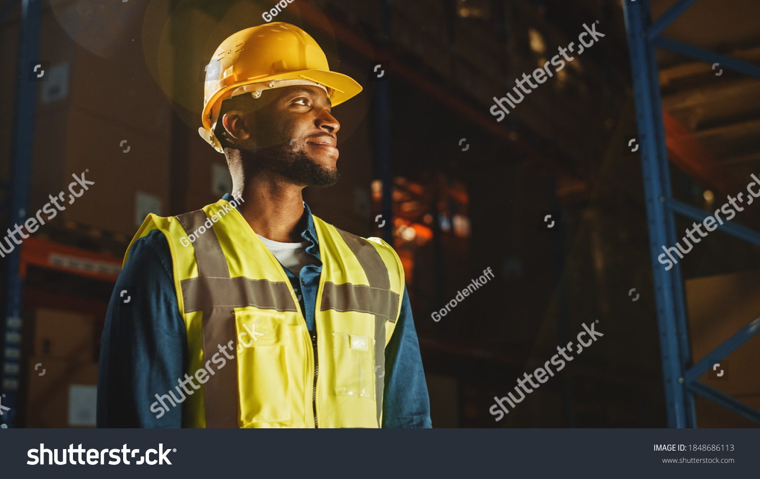 Handsome Professional Worker Wearing Safety Vest and Hard Hat Charmingly Smiling and Looks Into the Distance. In the Background Big Warehouse with Shelves full of Delivery Goods. Medium Portrait #1848686113