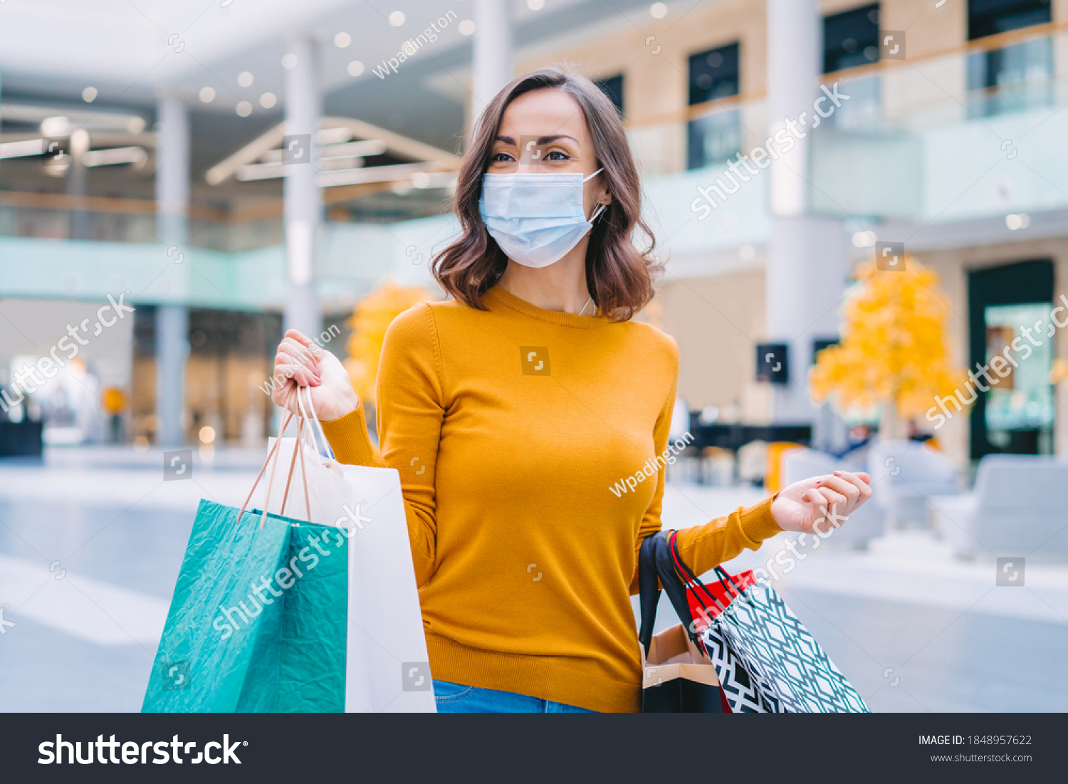 Portrait of casually dressed confident young woman wearing protecting medical mask while walking in mall with bunch of shopping bags in hands. Black friday sales concept. #1848957622