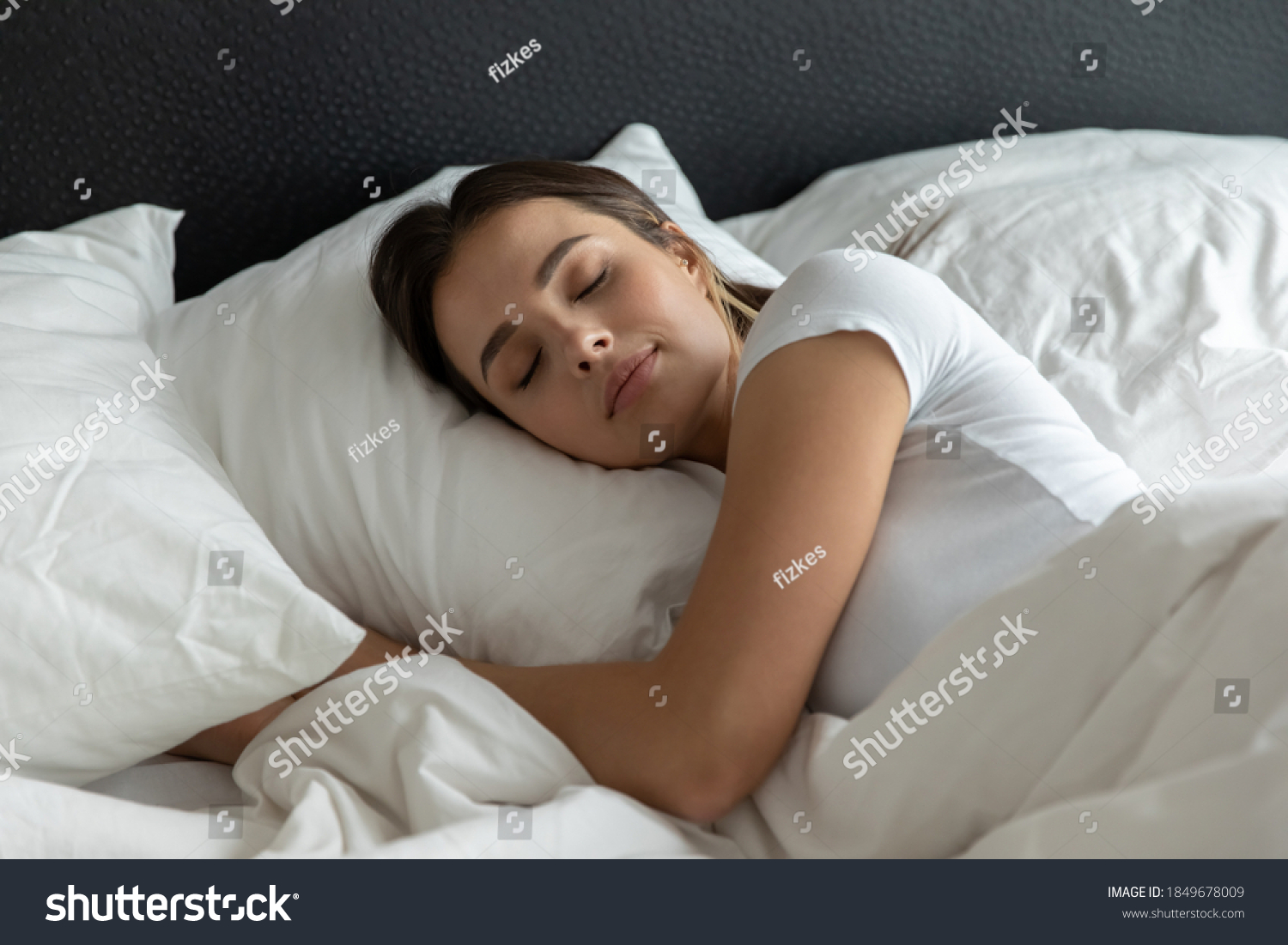 Sweet dreams. Relaxed young female sleeping in large cozy bed at home or in hotel suite room on clean white bedclothing hugging soft pillow covered with warm blanket dreaming resting having pleasure #1849678009