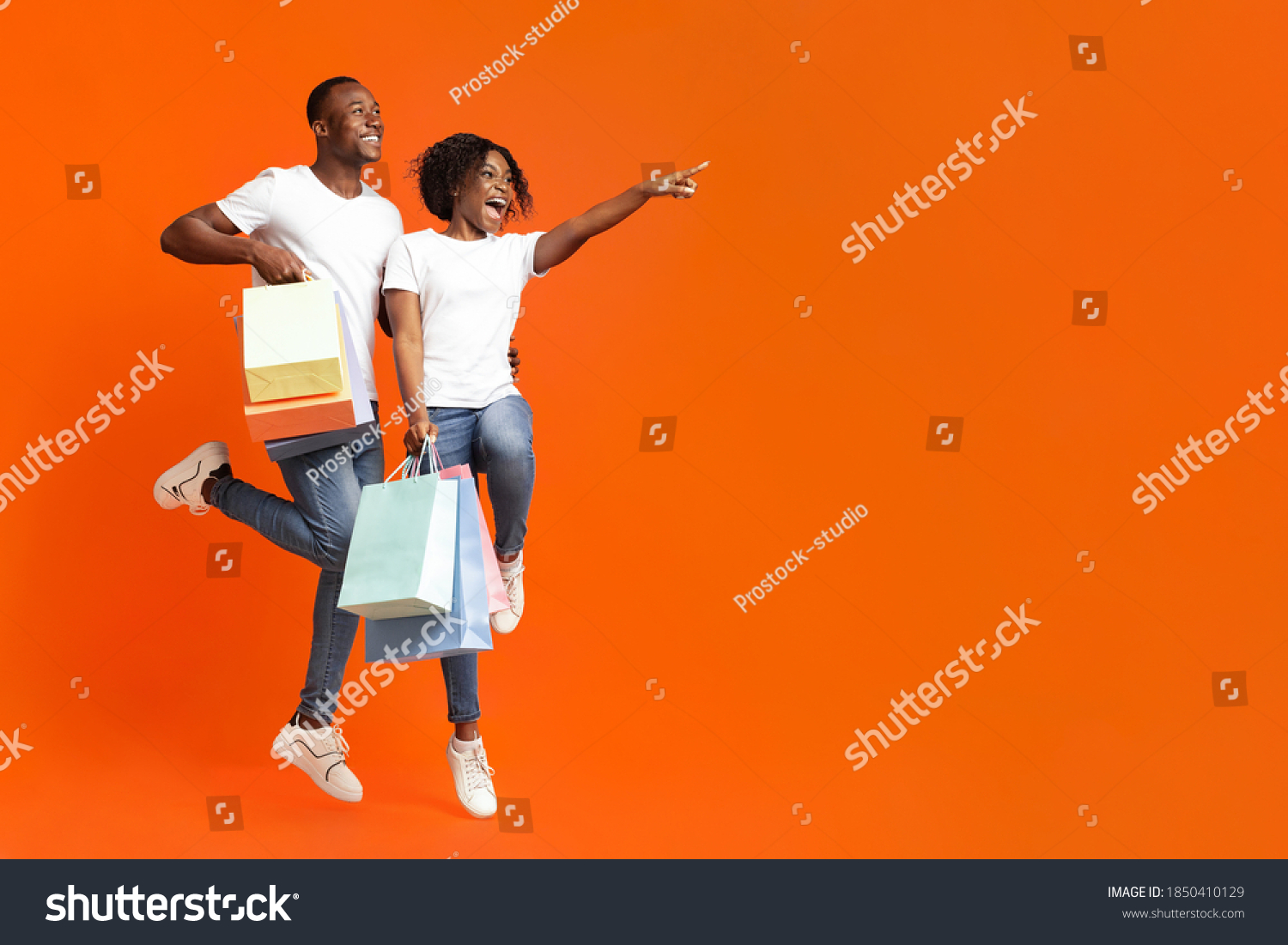 Emotional african-american couple with purchases jumping up and cheerfully pointing away at empty space, orange studio background. Happy young black man and woman aiming at advertisement #1850410129