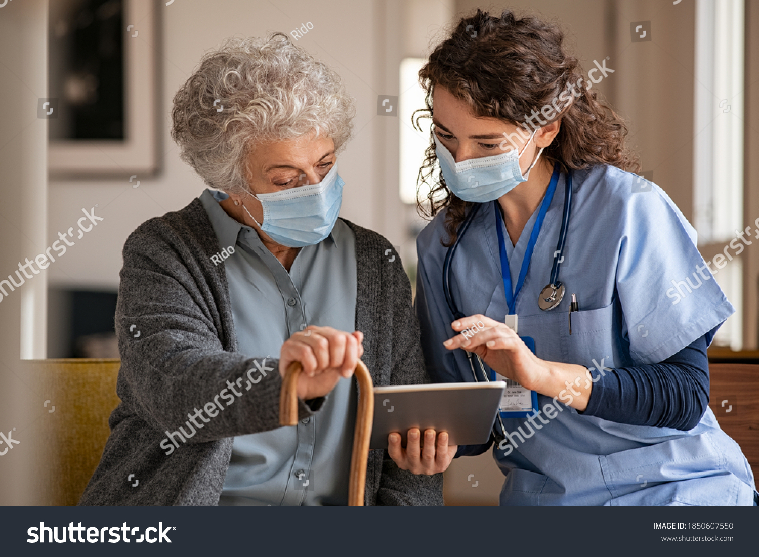 Doctor and senior woman going through medical record on digital tablet during home visit wearing face mask. Old woman with nurse with surgical mask and using digital tablet during coronavirus pandemic #1850607550