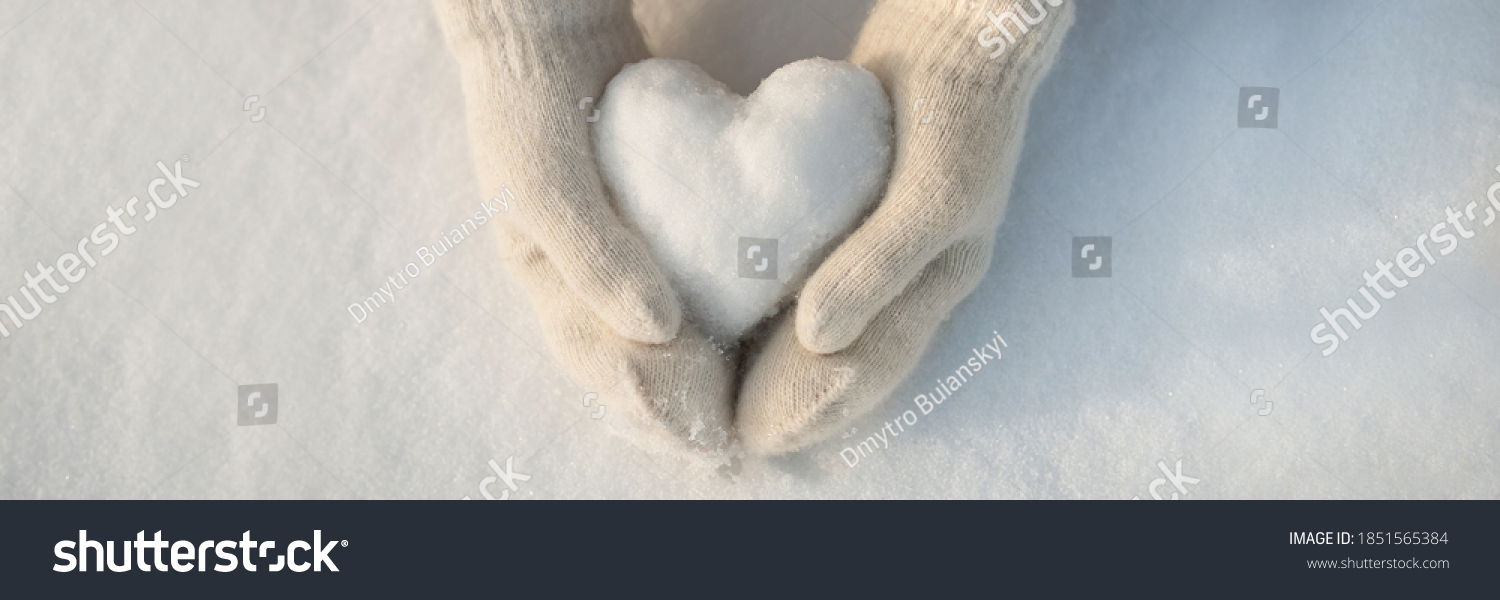 Snow heart in hands. Human hands in warm beige gloves with snowy heart against snow background. I love winter or St.Valentine's Day romantic creative concept. Panorama banner with free space for text #1851565384