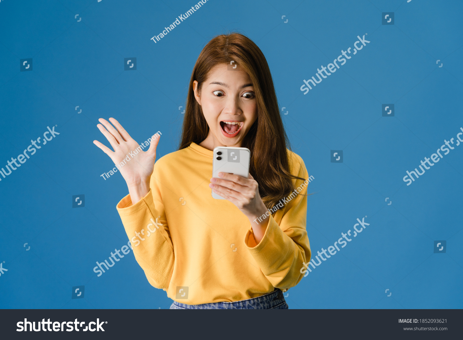 Surprised young Asia lady using mobile phone with positive expression, smiles broadly, dressed in casual clothing and standing isolated on blue background. Happy adorable glad woman rejoices success. #1852093621