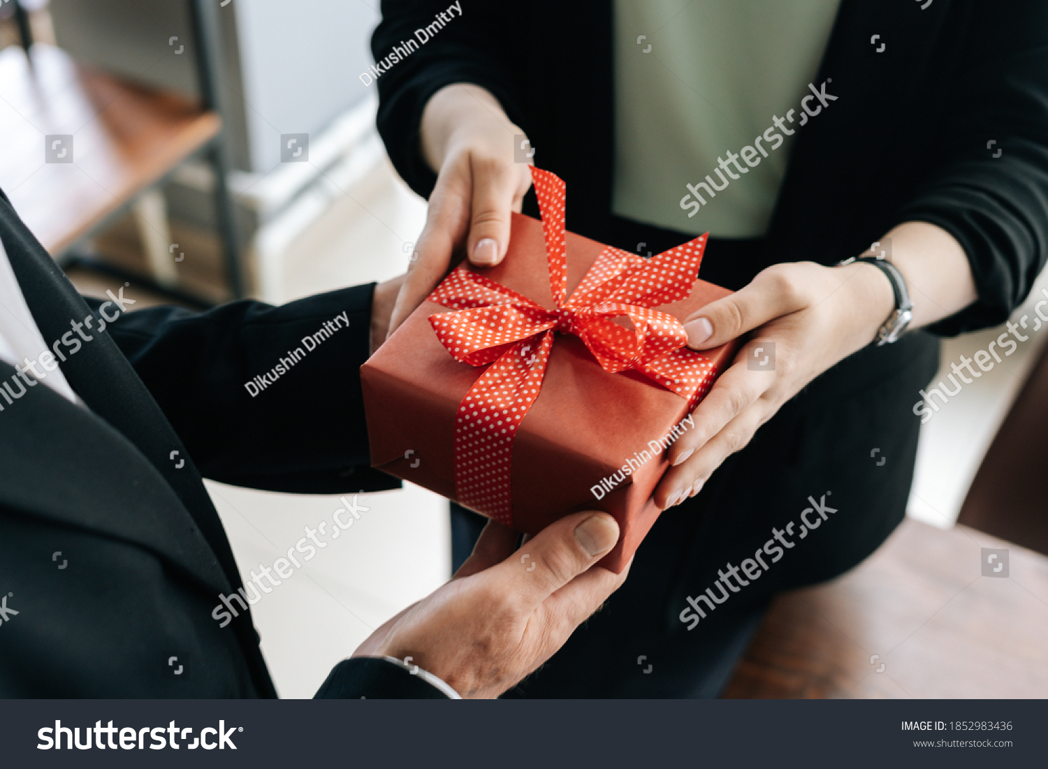Close-up view of hands of unrecognizable woman giving red gift box tied to bow handed to man. Giving gifts during the Christmas, Happy New Year and Happy Birthday at office. #1852983436