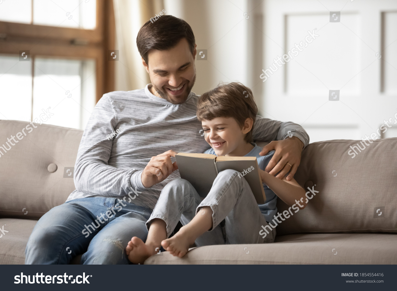 Loving happy father teaching adorable son to read, smiling dad and little boy child hugging, sitting on couch, holding book with fairy tale story, family spending weekend together, leisure time #1854554416