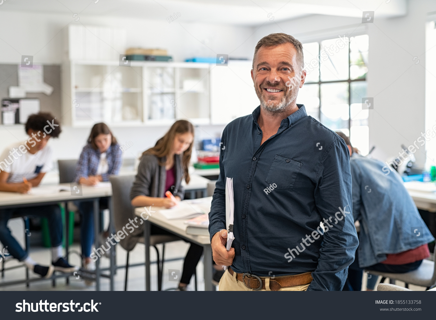 Portrait of mature teacher looking at camera with copy space. Happy mid adult lecturer at classroom standing after giving lecture. Satisfied high school teacher smiling while his students studying. #1855133758