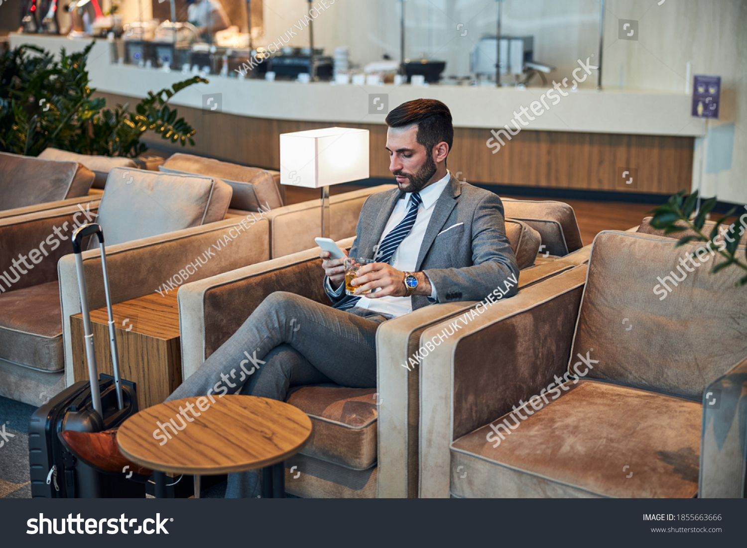 Businessperson waiting for his flight in an armchair while holding a glass of alcohol and browsing the smartphone #1855663666