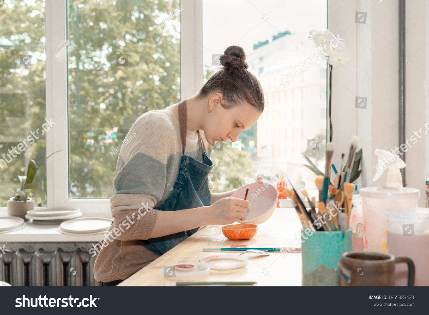 Skilled young woman in apron sitting at table and drawing on ceramic bowl in pottery workshop. Earn extra money, side hustle, turning hobbies into job. #1855983424