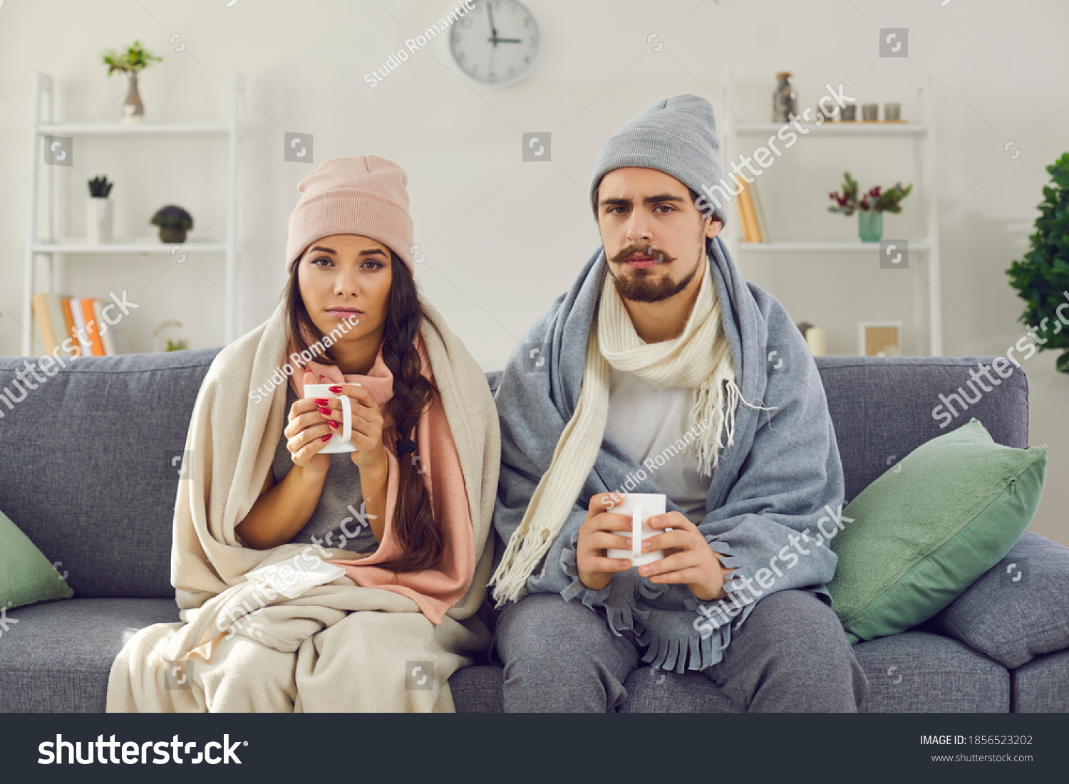 Dissatisfied young couple having problem with central heating, sitting on sofa at home, freezing, drinking hot tea trying to warm up. Sick man and woman wrapped in blankets suffering from cold or flu #1856523202