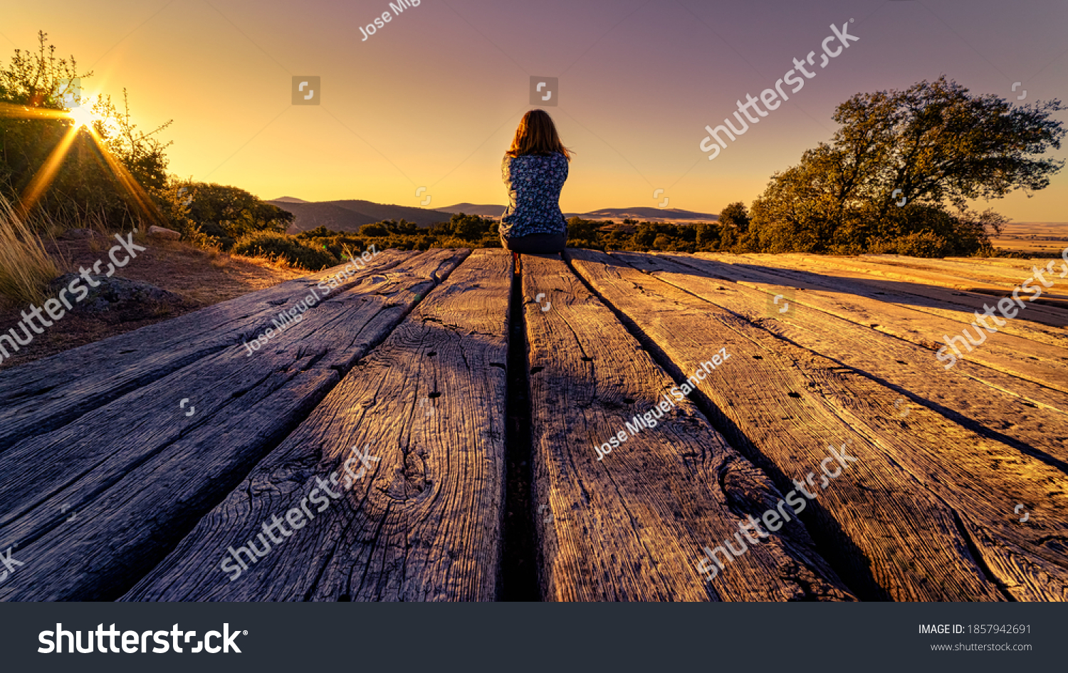 105 / 5000
Woman from behind sitting on a wooden floor, contemplating the horizon with sunset in the field. #1857942691