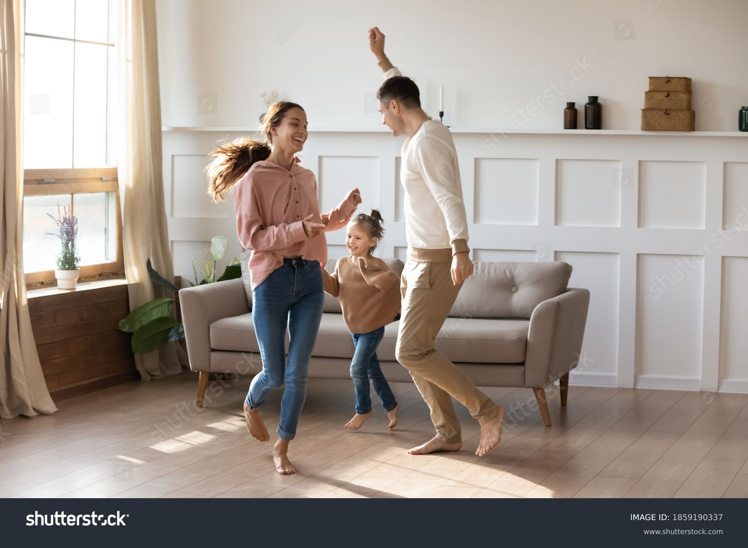 Cheery couple dancing with little daughter barefoot on wooden laminate floor with underfloor heating system in modern warm living room. New home, bank loan and lending, hobby and fun with kids concept #1859190337