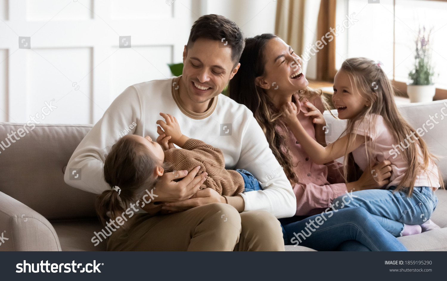 Funny parents play with two daughters resting on couch in light cozy living room. Family enjoy playtime together at home, tickling each other laughing feels happy. Having fun at home with kids concept #1859195290
