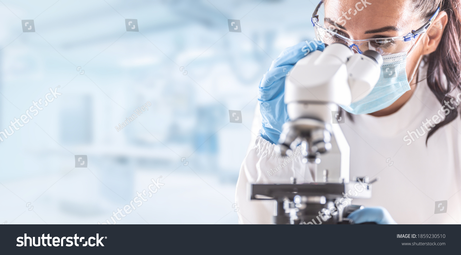 Female lab technician in protective glasses, gloves and face mask sits next to a microscope in laboratory. #1859230510