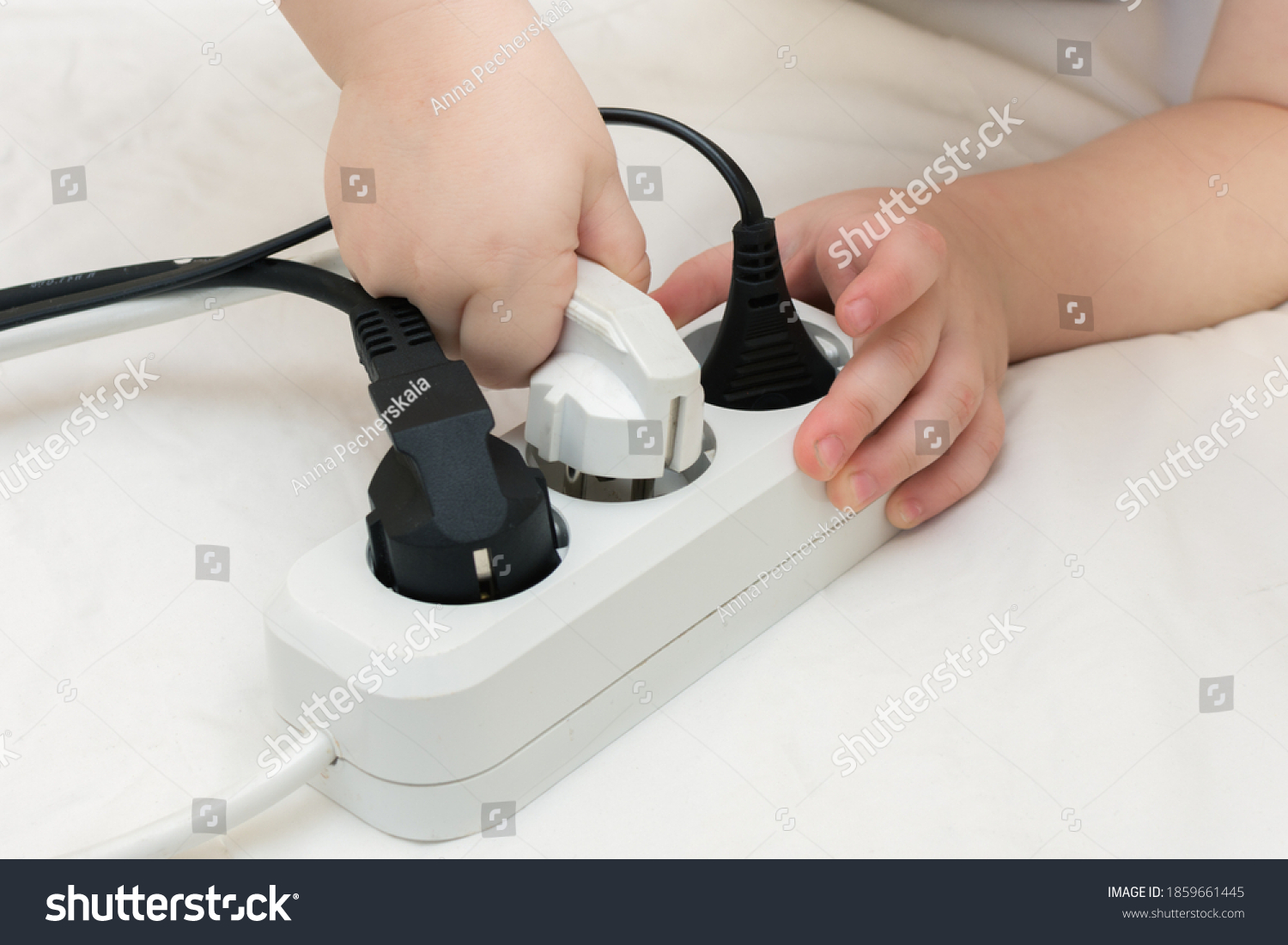Close-up of a child's hand inserting or removing a white electrical plug into a working outlet where there are already two black plugs. The idea is that playing with electricity can harm children #1859661445