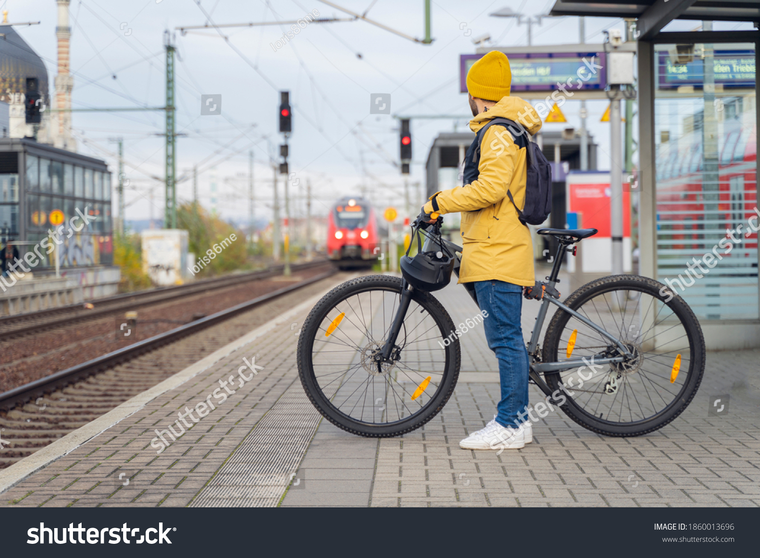 A man in winter clothes, waiting with his bike, the train. In the background the train that is about to arrive. #1860013696