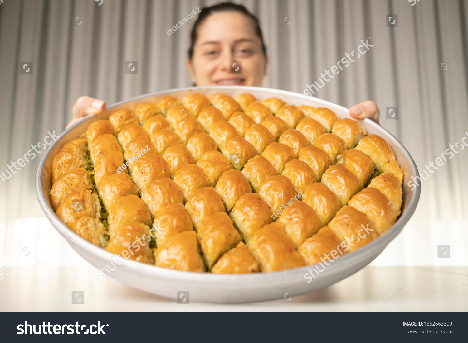 Close detail shot of a tray of crispy baklava dessert in the background smiling woman holding the tray in her hand #1862663809