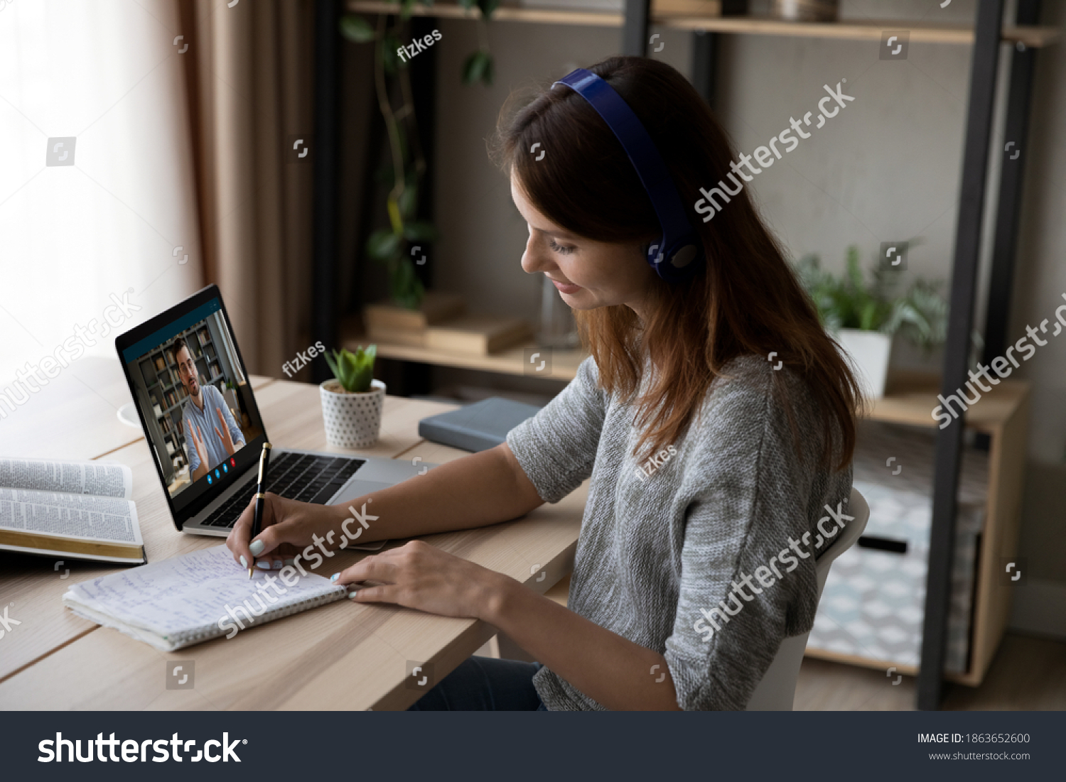 Side view smiling young woman in headphones holding video web camera call online meeting with male boss mentor, writing notes in copybook, happy lady practicing foreign language with native speaker. #1863652600