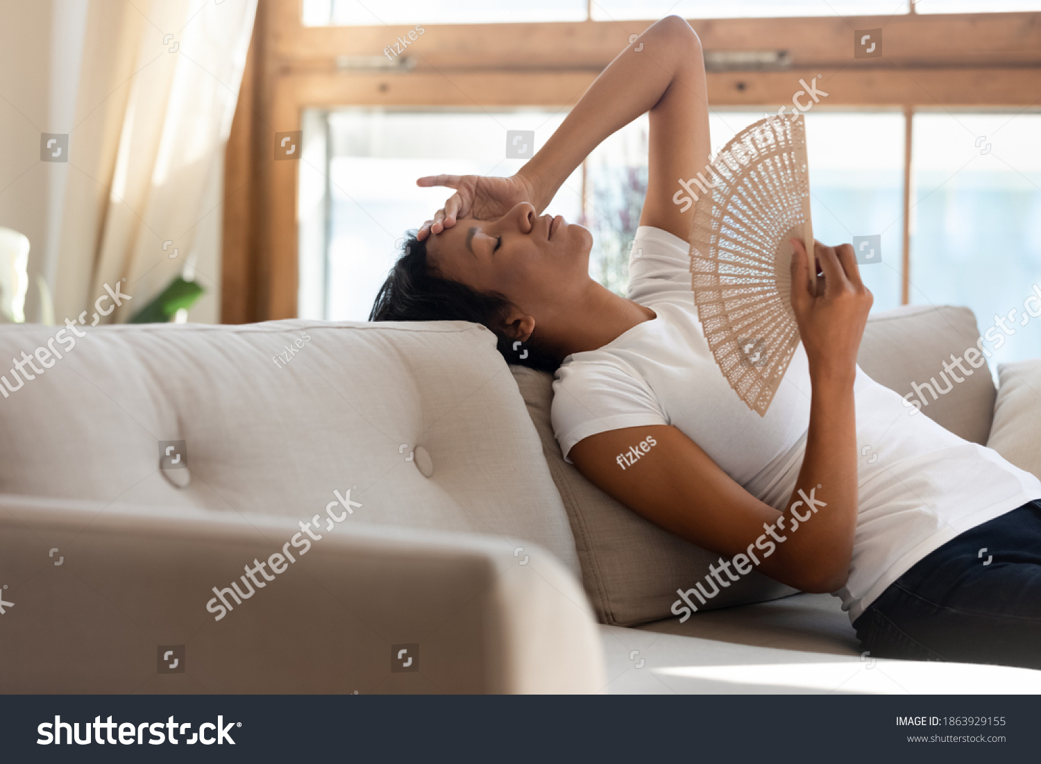 Tired unwell young African American woman sit lie on couch at home use waver suffer from hot weather indoors. Overheated millennial biracial female renter breathe fresh air from hand fan. #1863929155