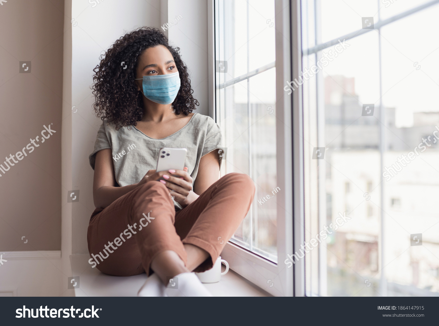 Sad woman alone during coronavirus pandemic wearing face mask indoors at home for social distancing. Mixed race girl looking at window. Anxiety, stress, lockdown, mental health crisis concept #1864147915