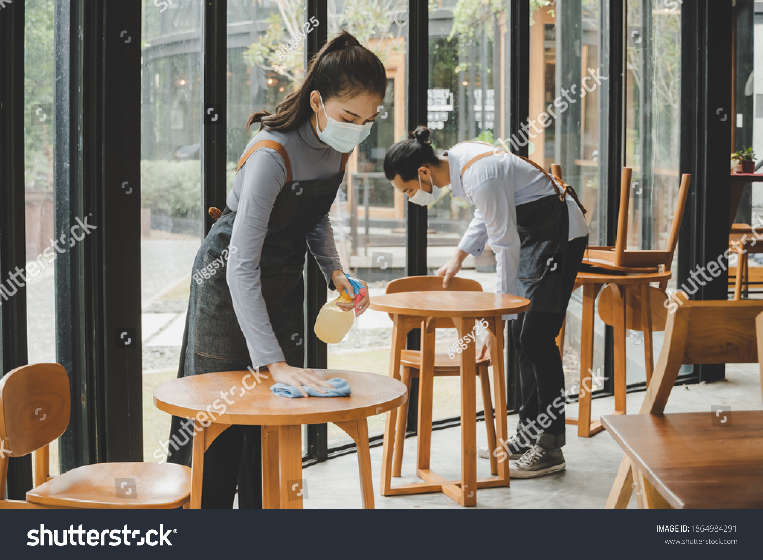 asian waitress staff wearing protection face mask in apron cleaning table with disinfectant spray for protect infection coronavirus (covid-19) in cafe coffee shop restaurant, hotel. new normal concept #1864984291