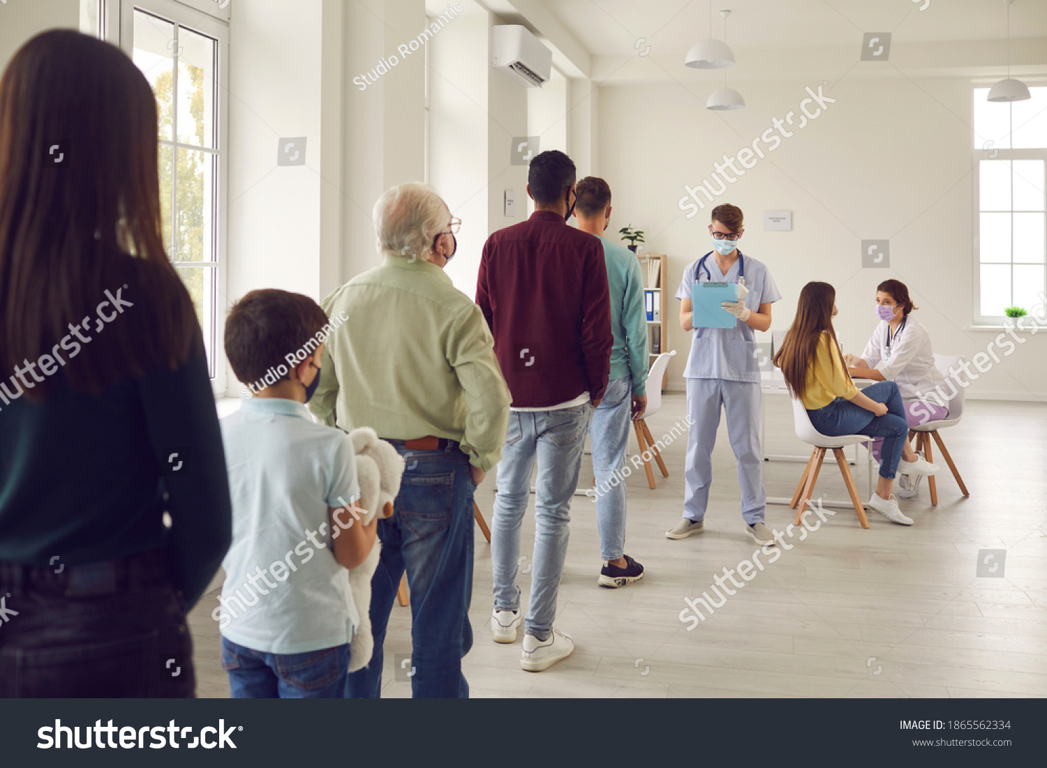 Diverse people lining up waiting for their turn to get shots at the hospital vaccination center. Young men and women, senior citizens and kids standing in queue for antiviral Covid-19 or flu vaccine #1865562334